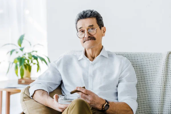 Homem sênior com bigode segurando sigar e cinzeiro nas mãos — Fotografia de Stock