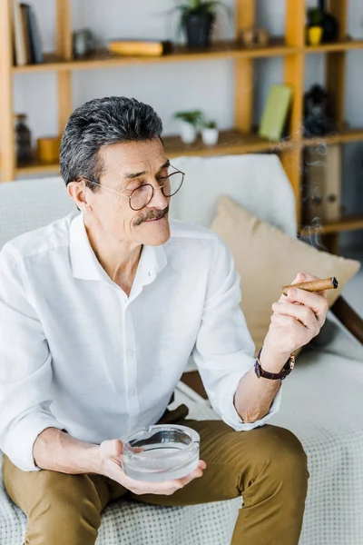 Uomo anziano allegro con baffi che tengono il sigaro e il posacenere in mani — Stock Photo
