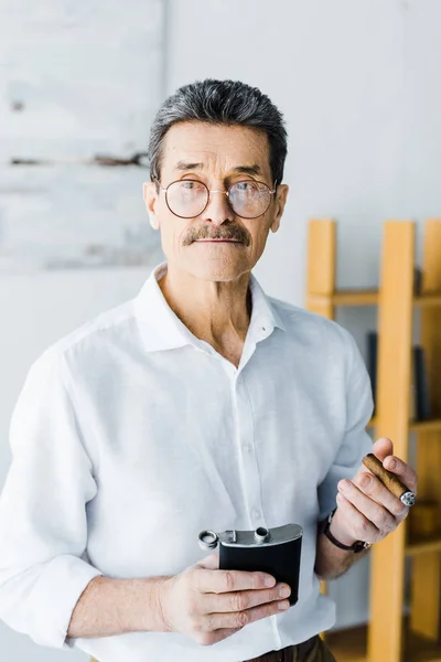 Пенсионер в очках держит в руках сигар и спиртовую фляжку — Stock Photo