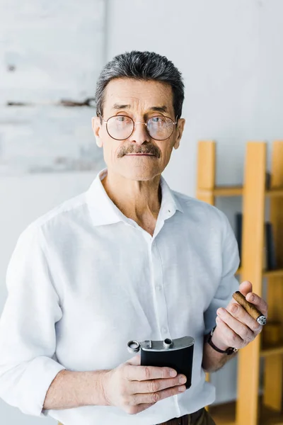 Retired man in glasses holding sigar and alcohol flask in hands — Stock Photo