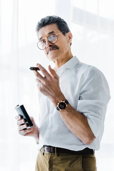 Pensioner in glasses holding sigar and alcohol flask in hands — Stock Photo