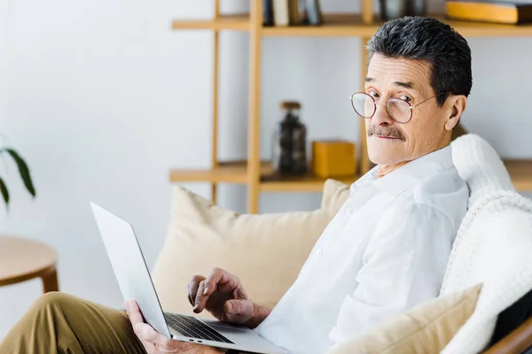 Sorprendido hombre mayor usando el ordenador portátil mientras está sentado en el sofá - foto de stock