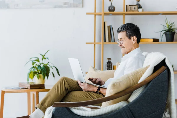 Senior homme dans des lunettes en utilisant un ordinateur portable tout en étant assis sur le canapé — Photo de stock