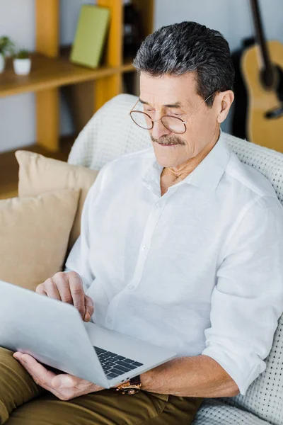 Retired man in glasses using laptop while sitting on sofa — Stock Photo