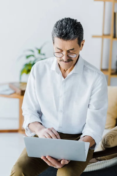 Pensioner with mustache and glasses using laptop while sitting on sofa — Stock Photo