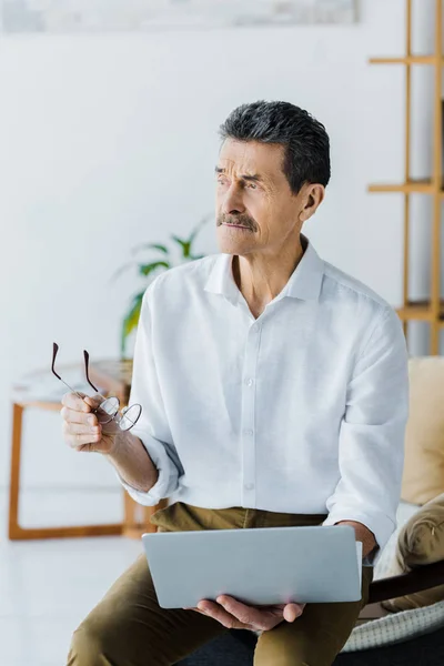 Pensionista pensativo com bigode segurando óculos e laptop em casa — Fotografia de Stock