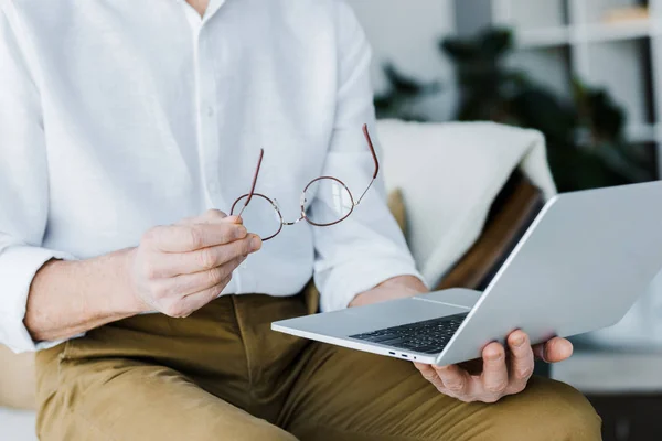 Обрезанный вид пенсионера, держащего очки и использующего ноутбук дома — стоковое фото