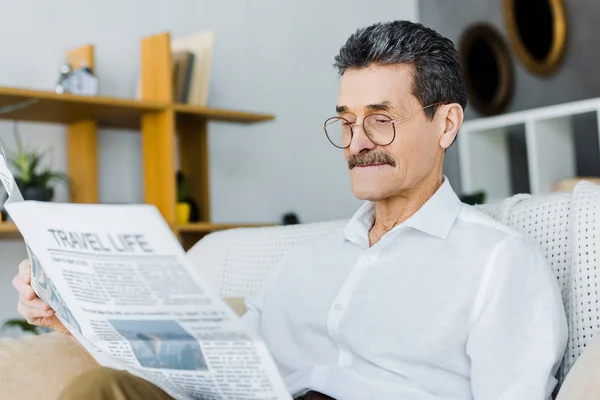 Cheerful senior man in glasses reading travel newspaper while sitting on sofa — Stock Photo