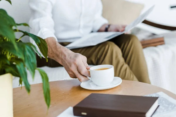 Cropped view of retired man holding cup with coffee and newspaper at home — Stock Photo