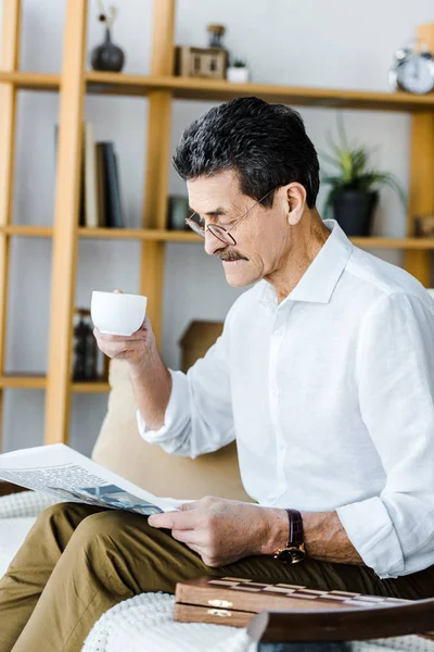 Pensionato in bicchieri in possesso di tazza con caffè e lettura giornale a casa — Foto stock