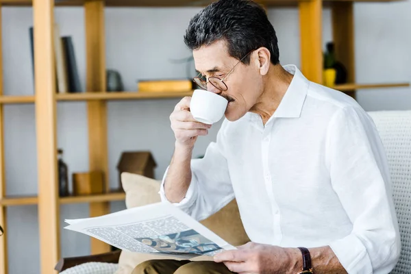 Pensioner in glasses drinking coffee and reading newspaper at home — Stock Photo