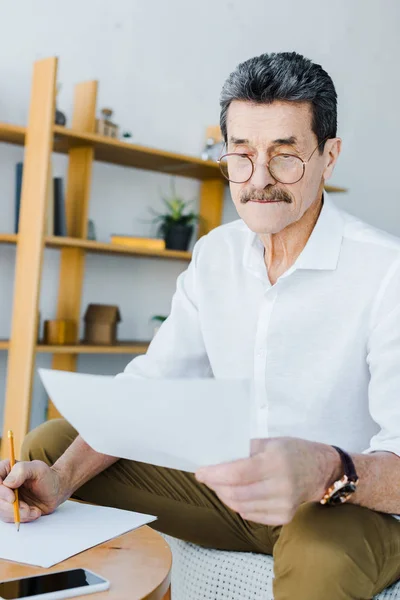 Senior mit Brille hält Bleistift in der Hand und schaut zu Hause auf Papier — Stockfoto