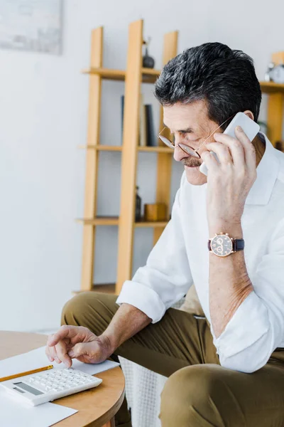 Uomo anziano parlando su smartphone e guardando calcolatrice a casa — Stock Photo