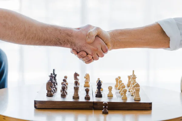 Vista recortada de los pensionistas estrechando la mano después de jugar al ajedrez en casa - foto de stock