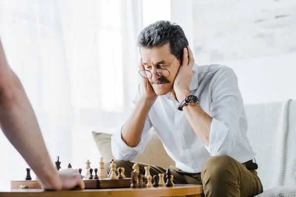 Selektiver Fokus eines älteren Mannes mit Schnurrbart, der die Ohren bedeckt, während er zu Hause mit einem Freund Schach spielt — Stockfoto