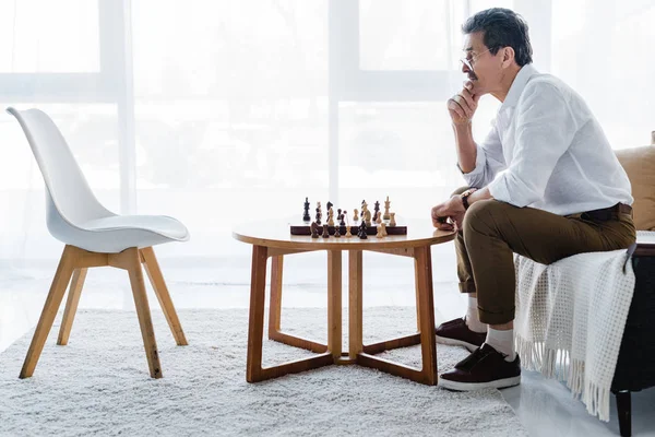 Pensativo hombre mayor con bigote mirando ajedrez en casa - foto de stock