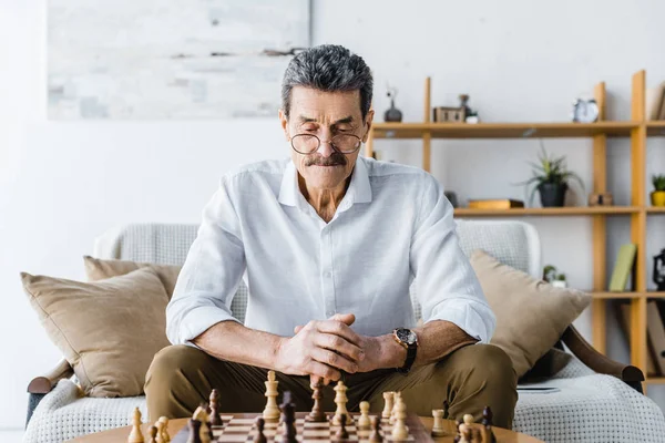 Pensativo hombre mayor con bigote mirando ajedrez en casa - foto de stock