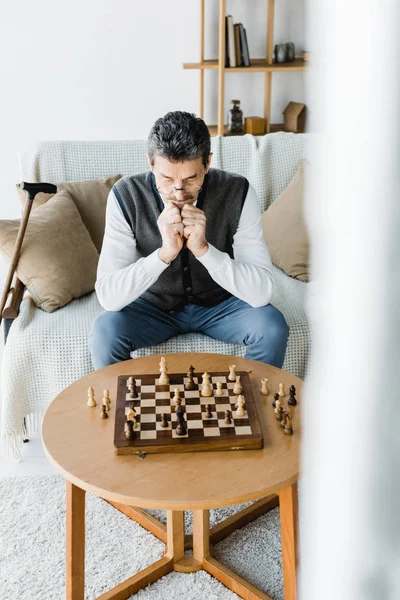 Pensionista reflexivo en gafas mirando tablero de ajedrez en casa - foto de stock