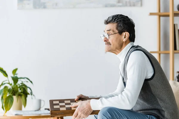 Pensativo pensionista em óculos segurando tabuleiro de xadrez em mãos — Fotografia de Stock