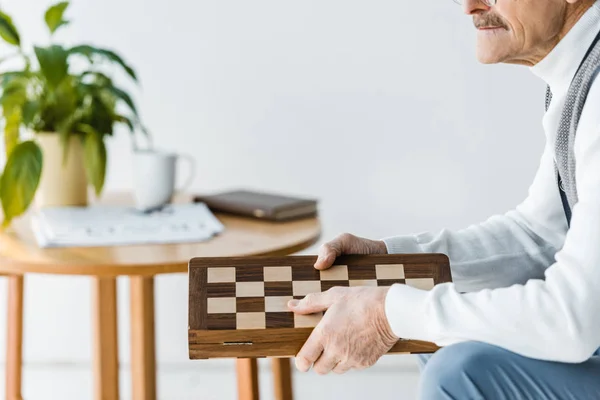 Ausgeschnittene Ansicht eines älteren Mannes, der zu Hause sitzt und ein Schachbrett hält — Stockfoto