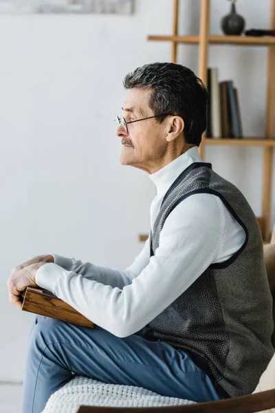 Retired man in glasses holding chess board at home — Stock Photo