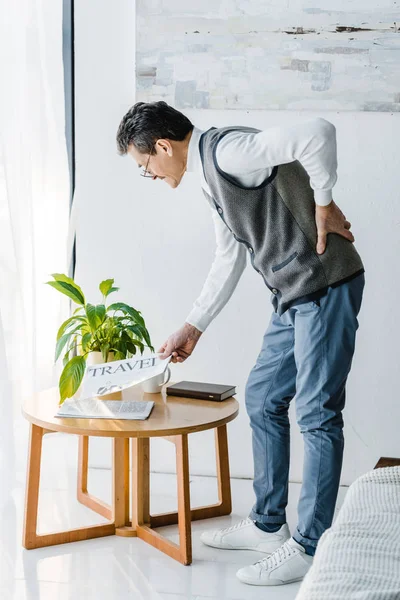 Senior hält sich zurück, während er Reisezeitung vom Kaffeetisch nimmt — Stockfoto