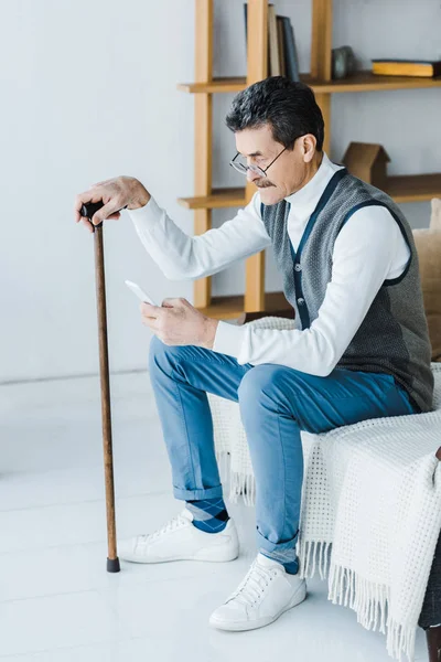 Homem sênior triste com bigode sentado no sofá e segurando smartphone em casa — Fotografia de Stock