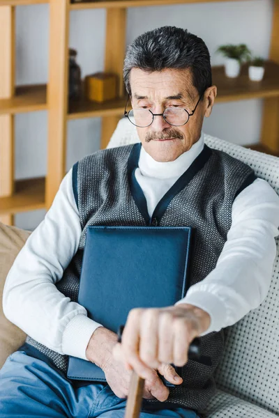 Aufgebrachter Senior mit Schnurrbart sitzt auf Sofa und hält Fotoalbum und Gehstock in der Hand — Stockfoto