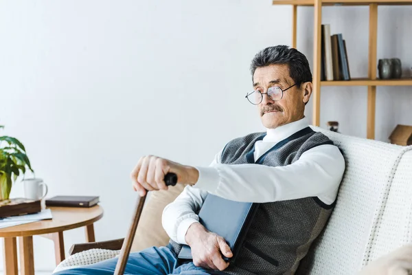 Retired man in glasses sitting on sofa with walking stick — Stock Photo