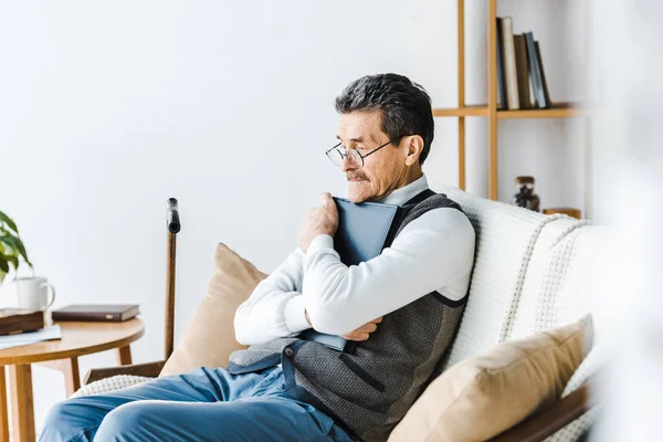 Hombre mayor en gafas abrazando álbum de fotos mientras está sentado en el sofá - foto de stock