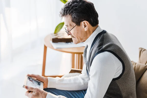 Trauriger Senior mit Brille betrachtet Foto im Fotorahmen, während er auf dem Sofa sitzt — Stockfoto