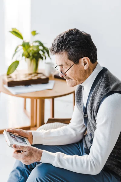 Upset senior man in glasses looking at photo in photo frame while sitting on sofa — Stock Photo