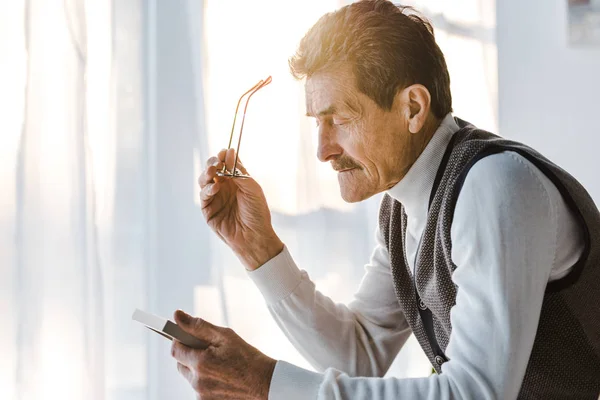 Sad pensioner holding glasses and looking at photo in photo frame while sitting at home — Stock Photo