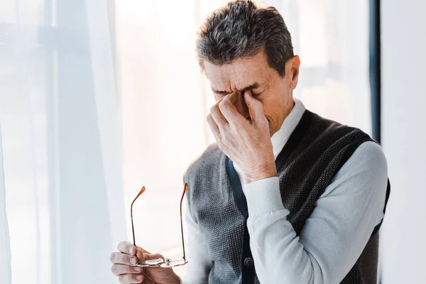 Homme âgé épuisé avec les cheveux gris touchant les yeux tout en tenant des lunettes — Photo de stock