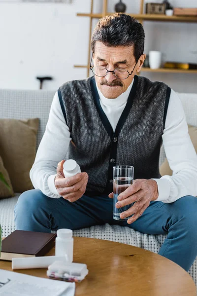 Hombre mayor mirando botella con pastillas mientras sostiene el vaso de agua - foto de stock