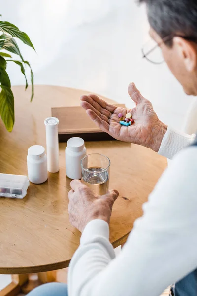 Selective focus of retired man looking at pills while holding glass of water — Stock Photo