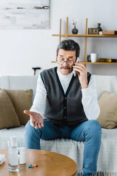 Selektiver Fokus eines älteren Mannes, der in der Nähe von Tabletten und Glas Wasser mit dem Smartphone spricht — Stockfoto