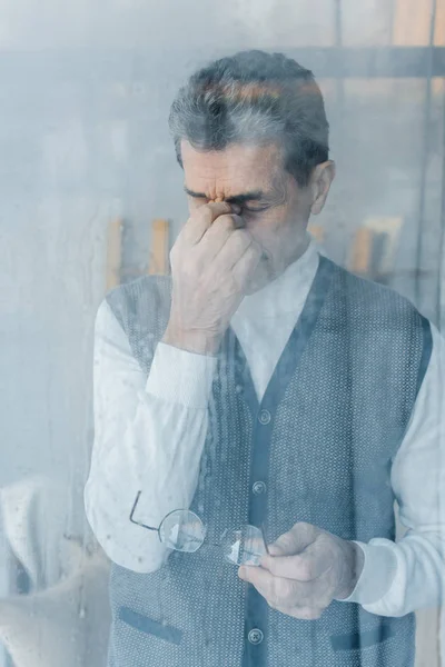 Tired senior man touching eyes while standing near window — Stock Photo