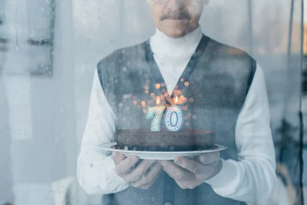 Recortado vista de triste anciano sosteniendo pastel de cumpleaños cerca de ventana - foto de stock