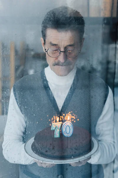 Uomo anziano solitario che tiene la torta di compleanno vicino alla finestra a casa — Foto stock
