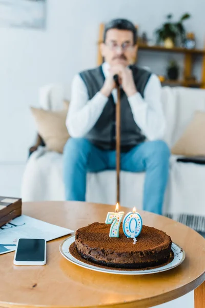 Selective focus of birthday cake with burning candles with sad senior man holding walking cane on background — Stock Photo