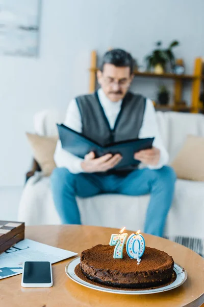 Foco seletivo de bolo de aniversário com velas em chamas com o homem sênior solitário segurando álbum de fotos no fundo — Fotografia de Stock