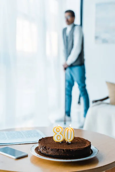 Foco seletivo de bolo de aniversário com velas em chamas com o homem aposentado de pé com bengala no fundo — Fotografia de Stock