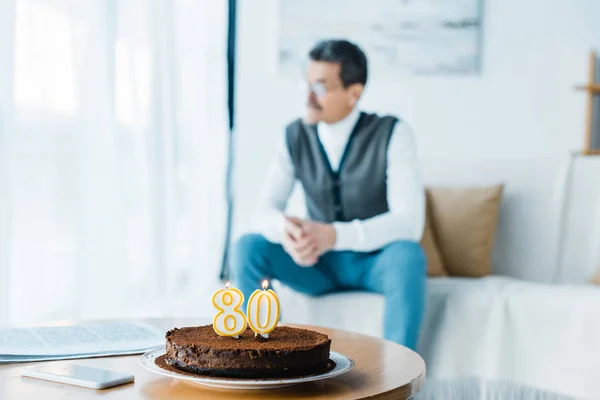 Foco seletivo de bolo de aniversário com velas acesas com o homem sênior solitário sentado no fundo — Fotografia de Stock