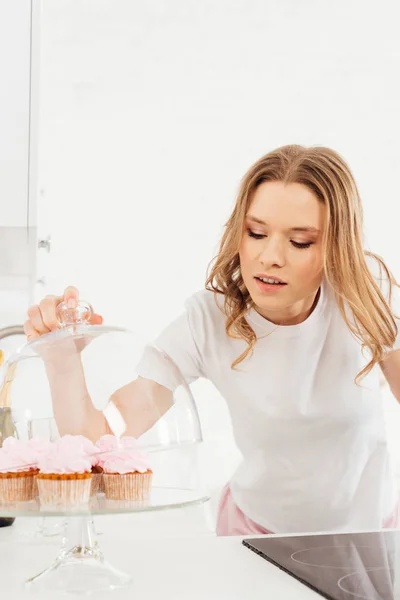 Beautiful girl in pajamas holding dome from glass stand with pink cupcakes in kitchen — Stock Photo