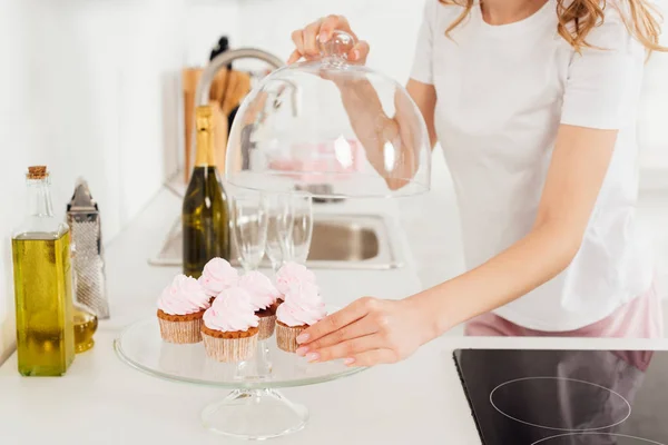 Cropped view of girl in pajamas holding dome from glass stand with pink cupcakes in kitchen — Stock Photo