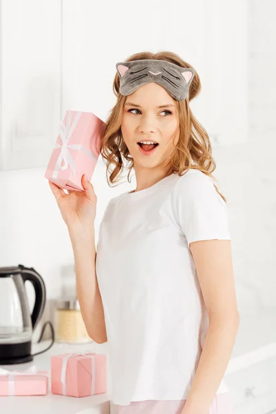 Красива здивована дівчина в піжамі і спальна маска, що тримає подарункову коробку на кухні — стокове фото