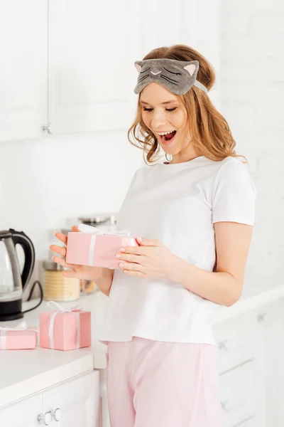 Beautiful surprised girl in pajamas and sleeping mask holding gift box in kitchen — Stock Photo