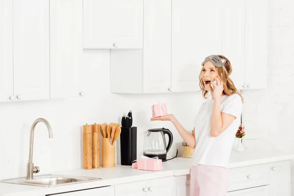 Hermosa chica emocionada en pijama y la máscara de dormir celebración de caja de regalo y hablar en el teléfono inteligente en la cocina - foto de stock