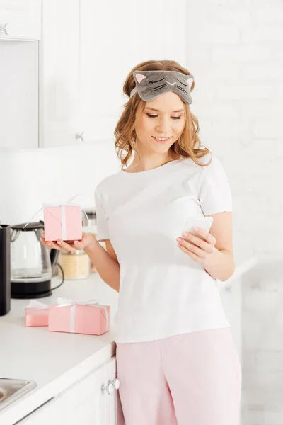 Красивая улыбающаяся девушка в пижаме и маске для сна, держащая подарочную коробку и используя смартфон на кухне — стоковое фото